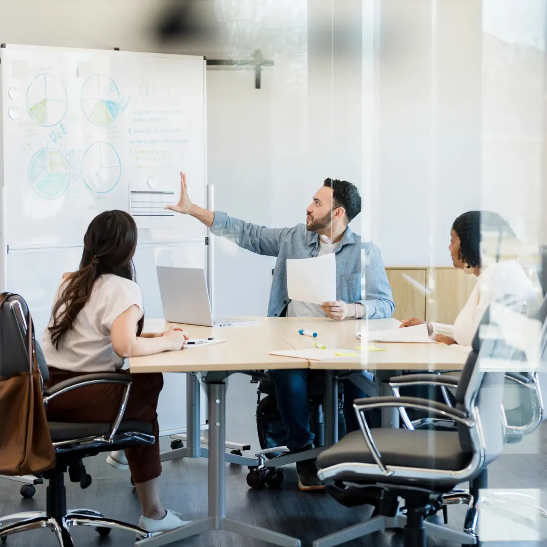 three people sitting in a board room with one person pointing to a whiteboard